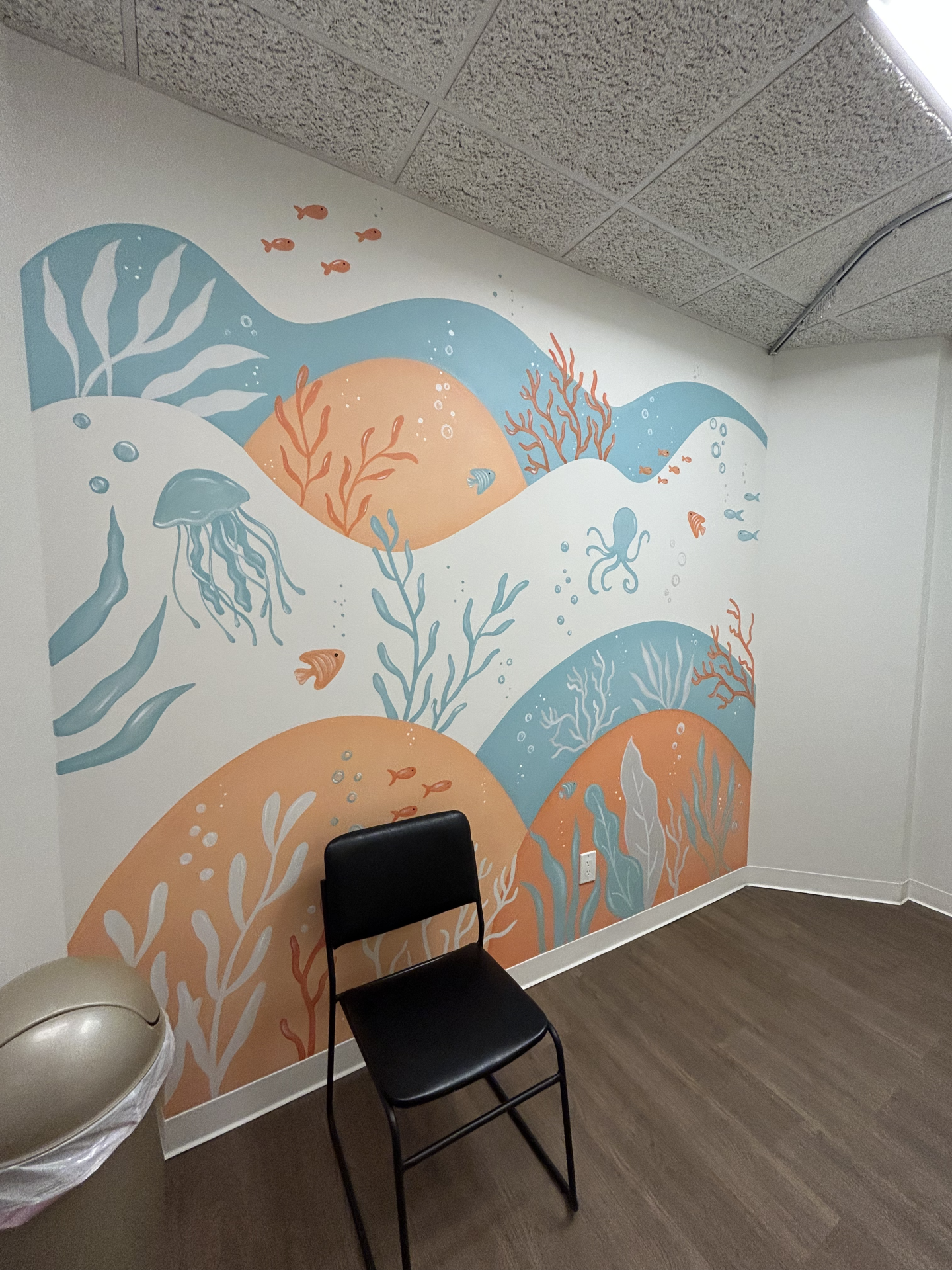 A mural on a white wall featuring abstract blue and orange waves, sea creatures including an octopus and jellyfish, and seaweed, with a black chair positioned in front.