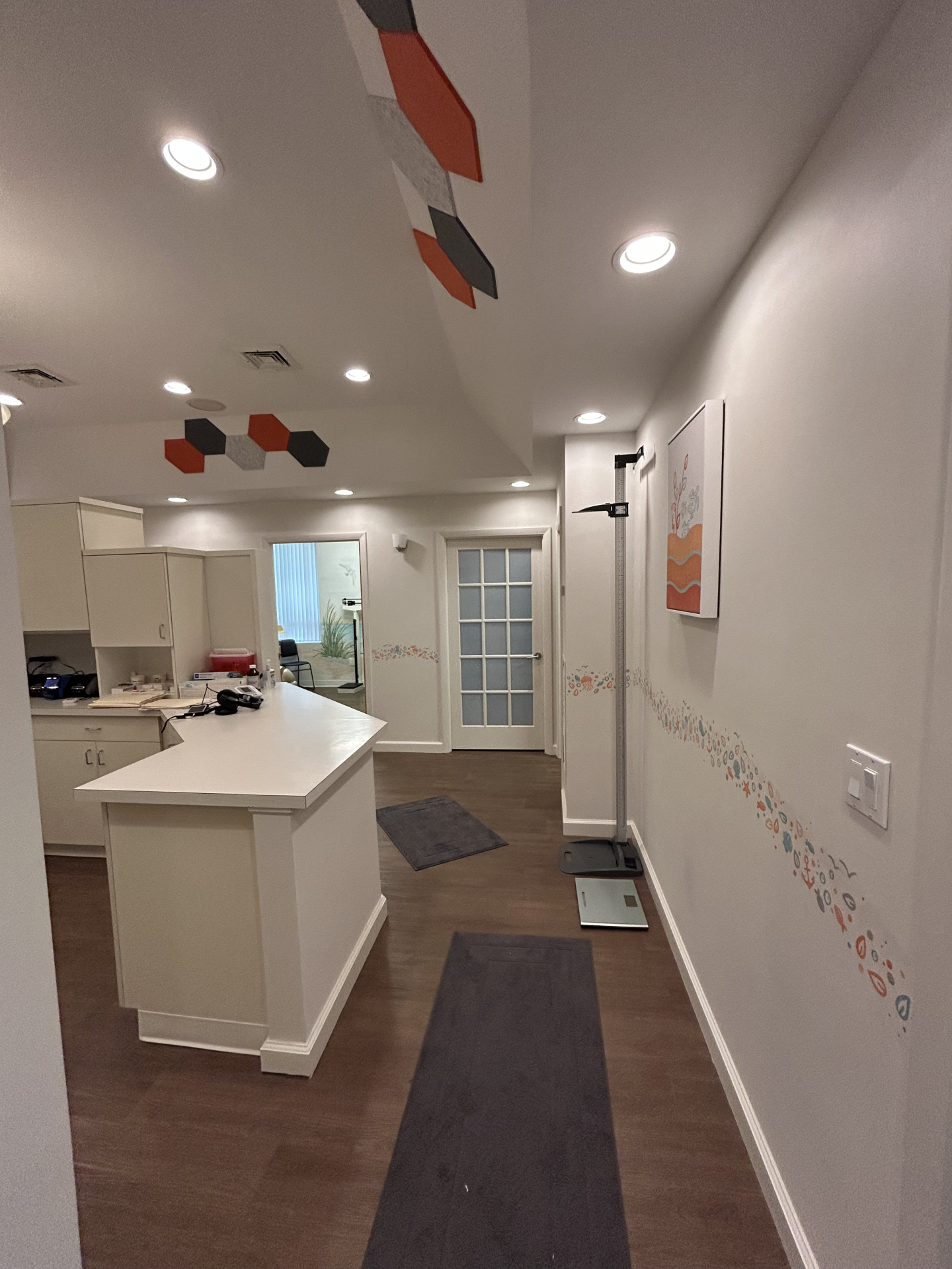 A room with wood-look flooring, a central desk, hexagonal wall accents, and a framed abstract painting on the ceiling.