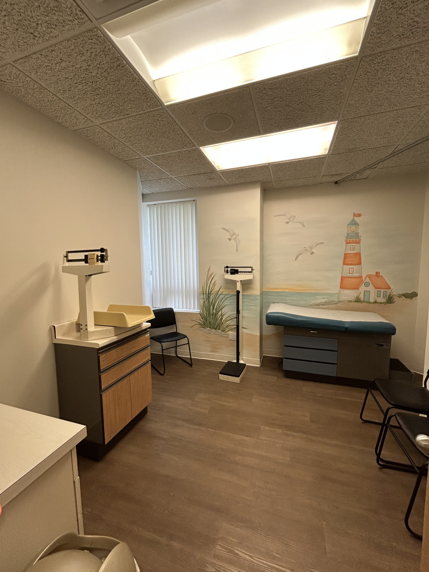 Exam room with a medical scale, an examination table, and a wall mural featuring a lighthouse.