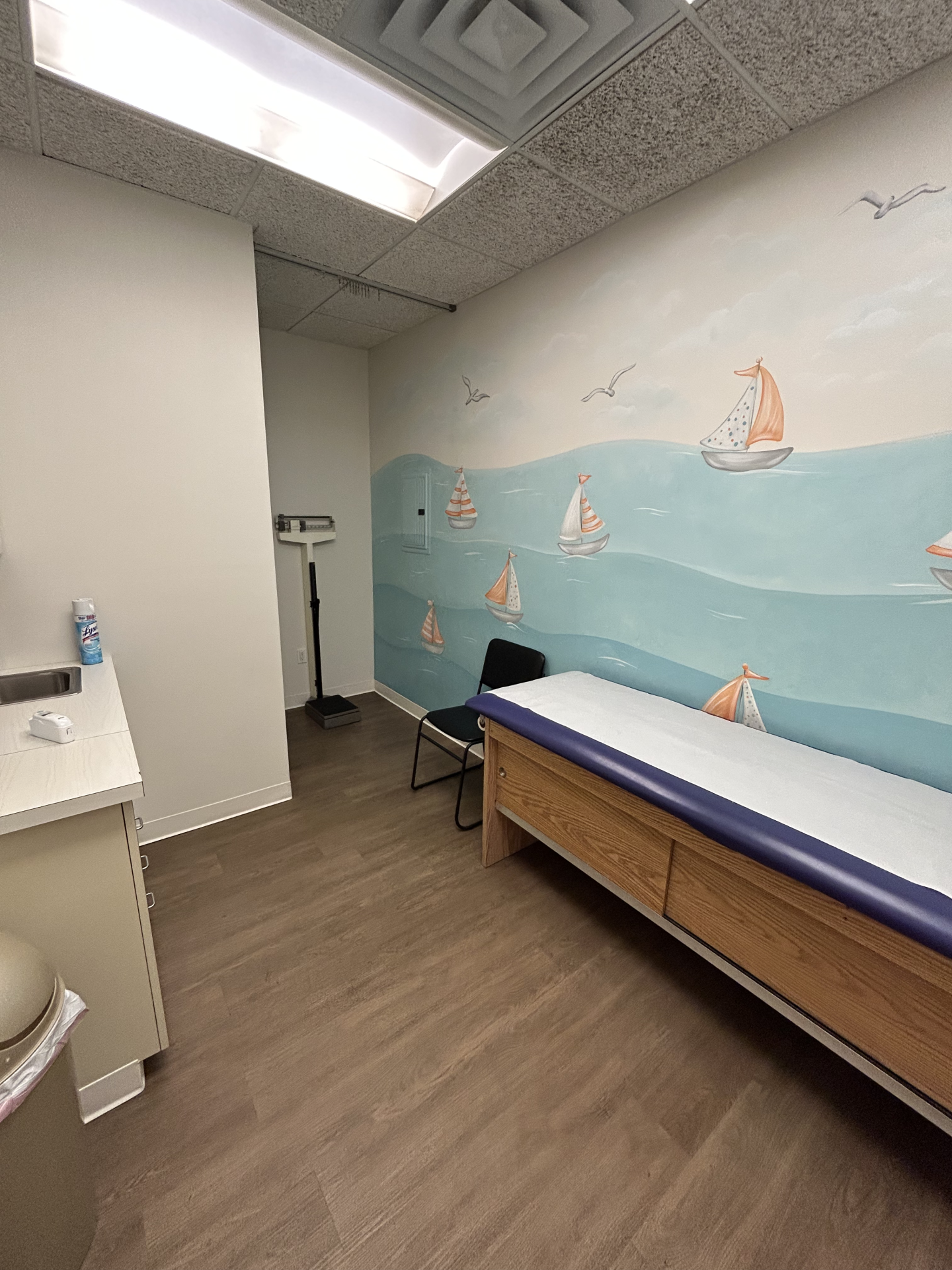 A pediatric exam room featuring a wall mural of sailboats on the ocean, an exam table with a blue pad, and a medical scale.
