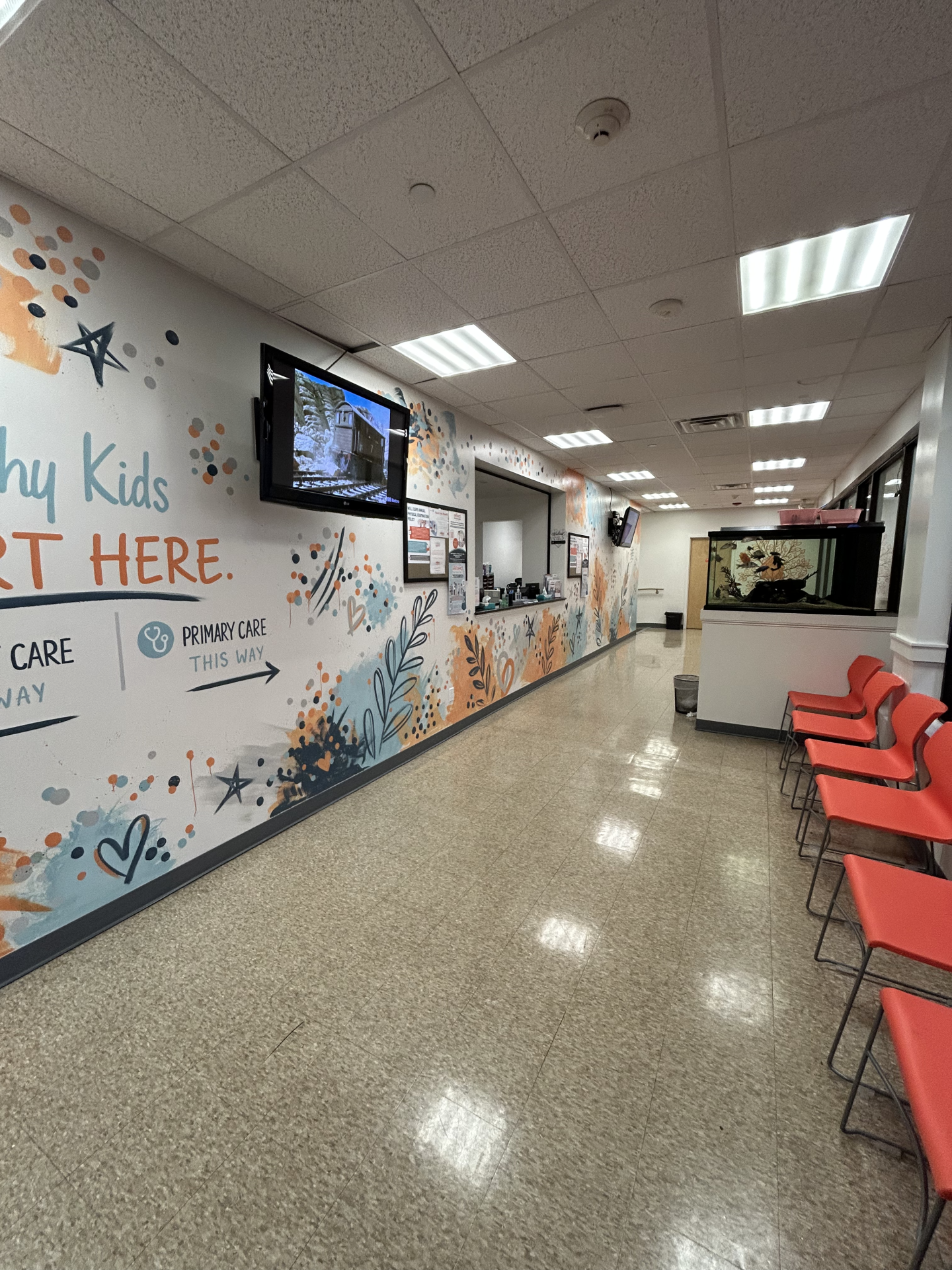 A waiting room featuring a mural with text that reads Primary Care This Way and chairs lined up along the side wall.