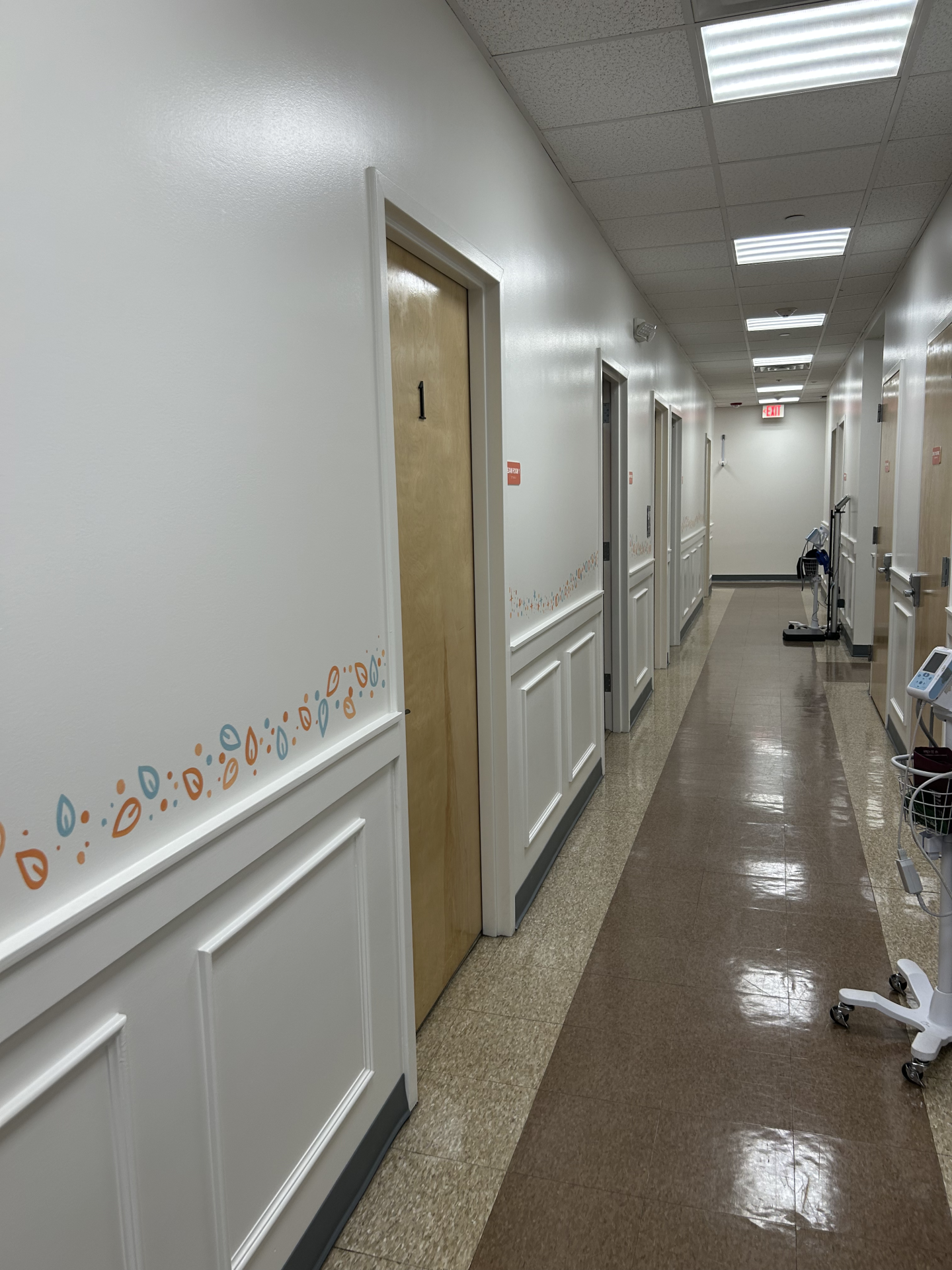 A long hospital hallway with white doors, colorful leaf-patterned wall decals, and a red exit sign at the far end.
