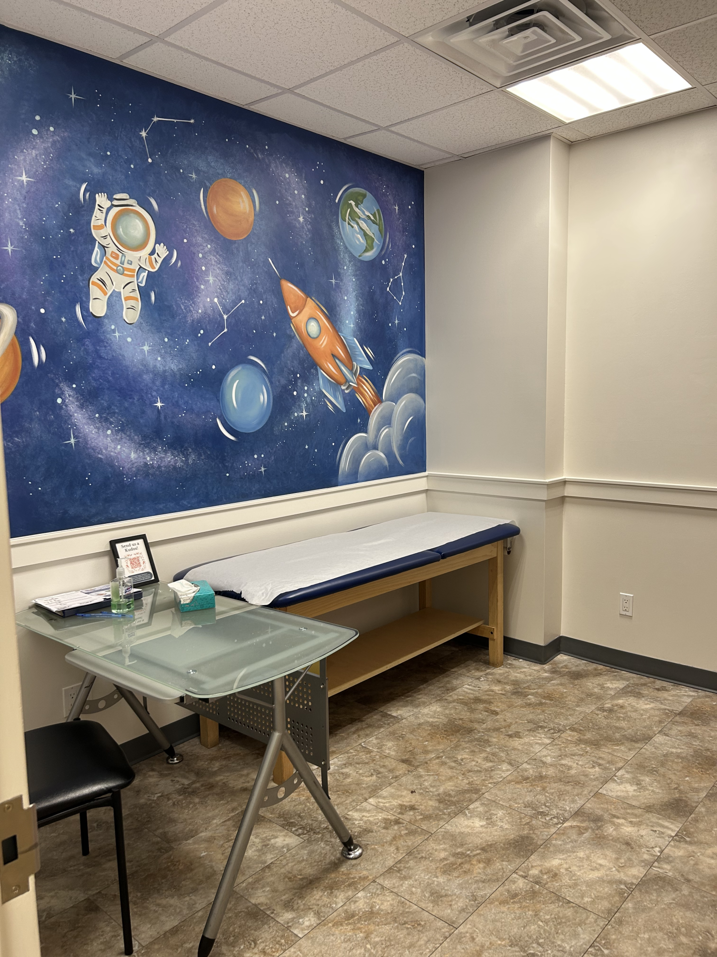 A medical examination room with a space-themed mural on the wall, an exam table, and a glass-topped desk.