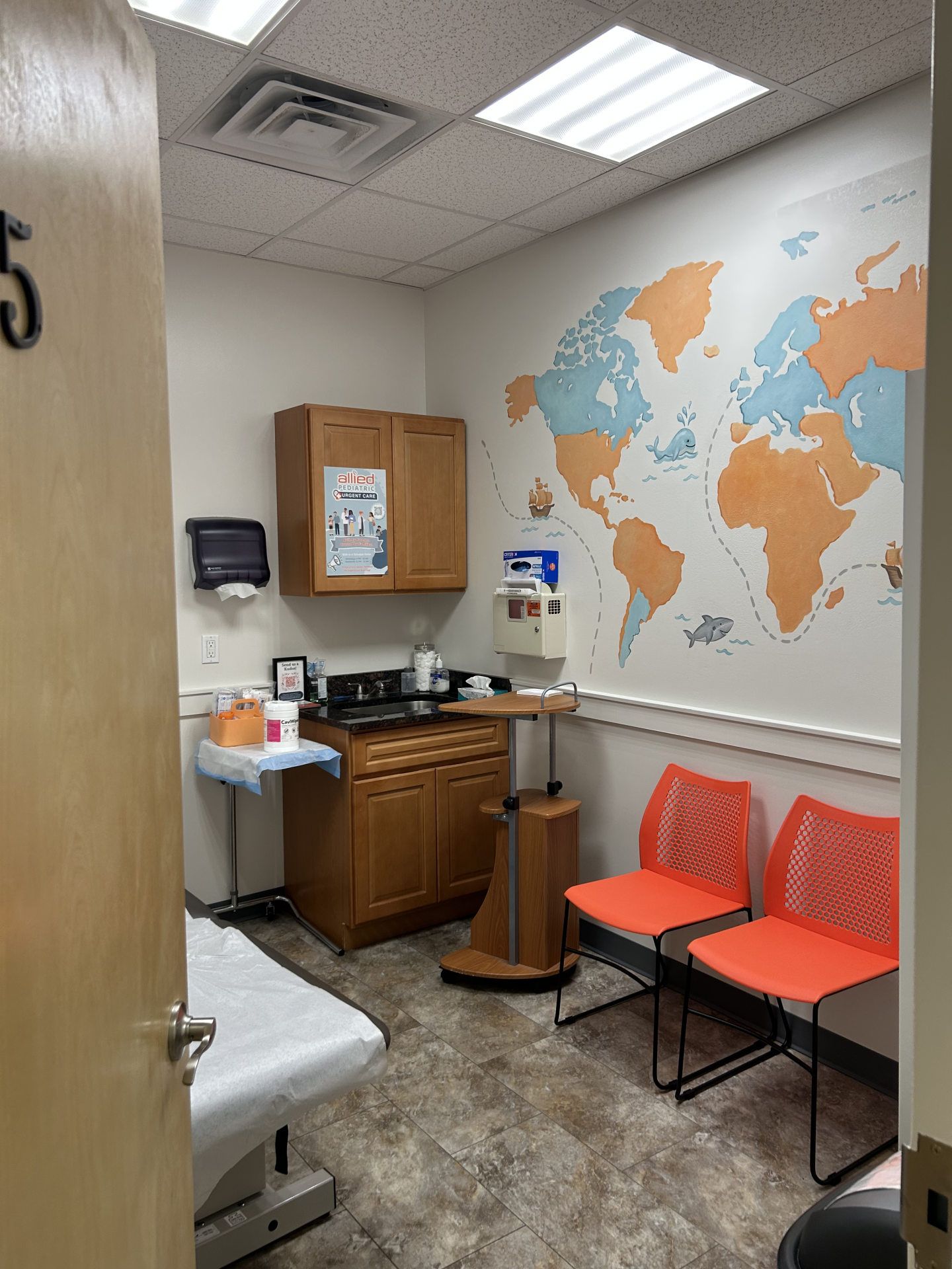 A pediatric exam room featuring a wall-mounted map mural with cartoon sea creatures, two orange chairs, a wooden cabinet with a poster, and a medical exam table.