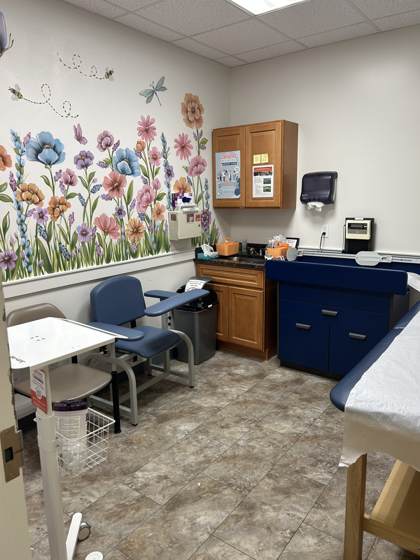 A medical examination room with a floral wall mural, blue cabinets, and a grey exam table.