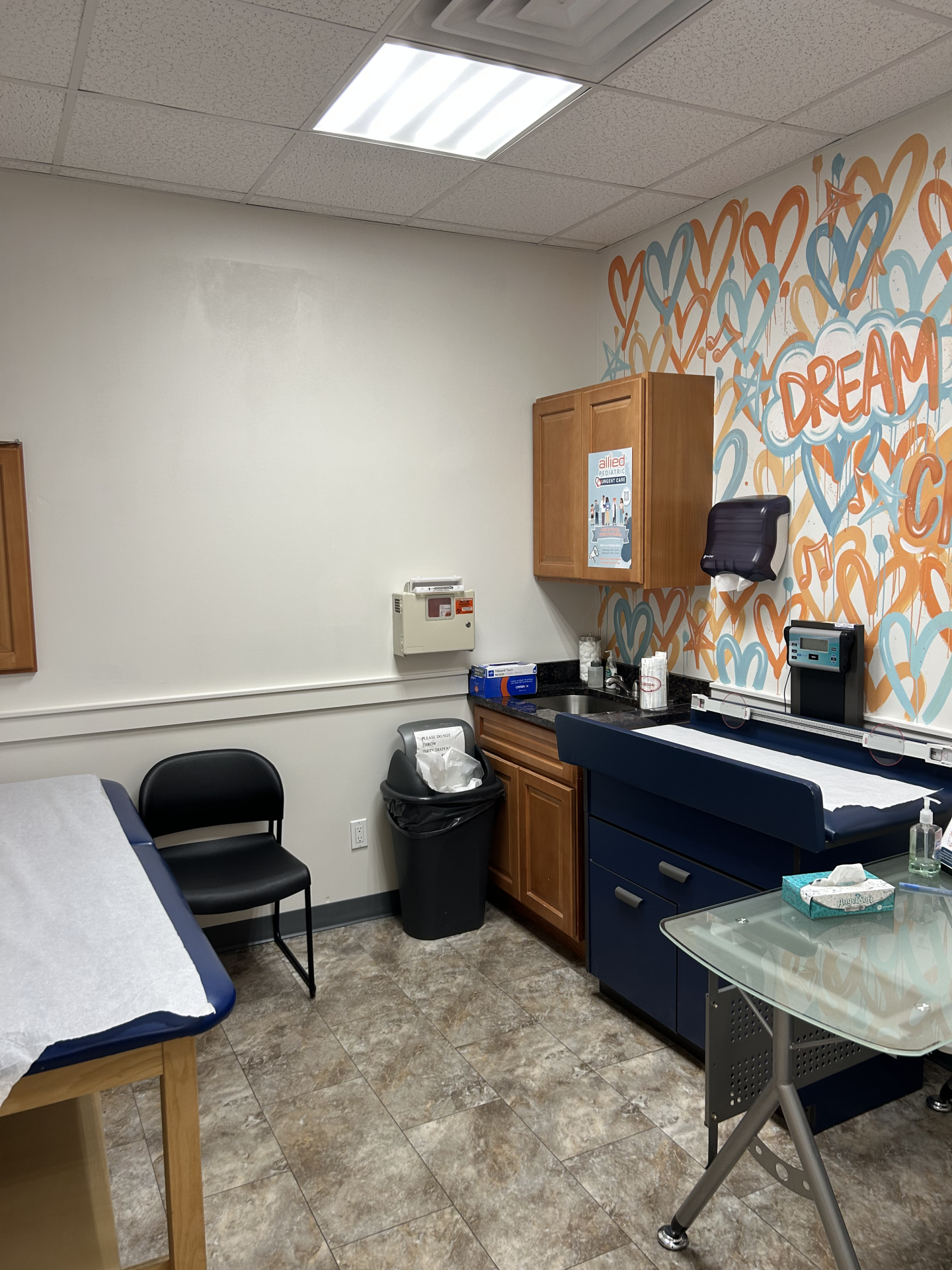 An exam room with a wooden cabinet, a blue treatment table, a rolling glass cart, and a wall featuring colorful graffiti-style text.