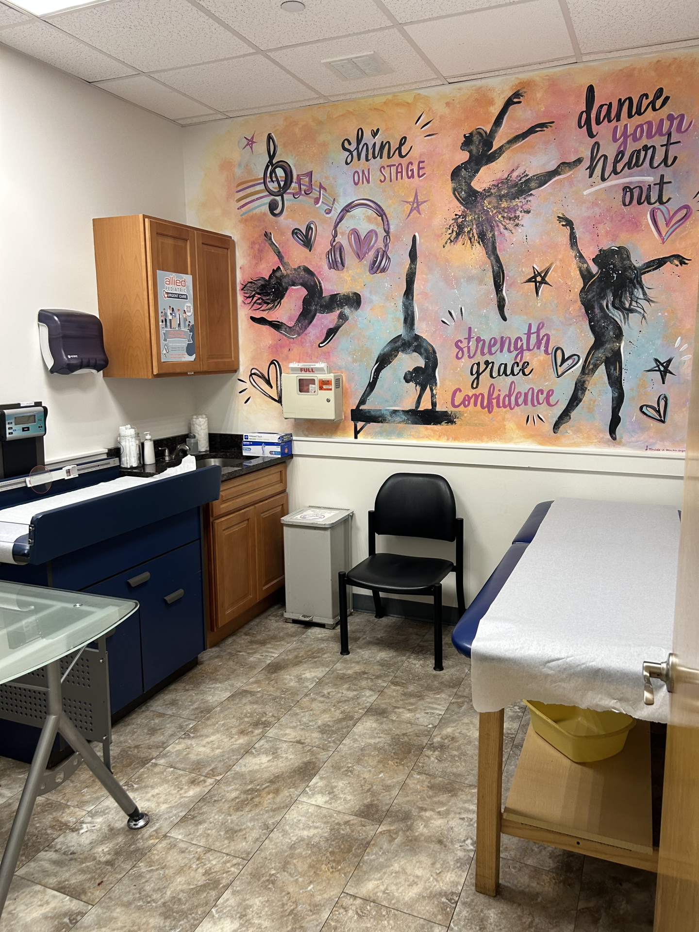 Exam room with an exam table, a black chair, and a wall mural featuring silhouettes of dancers and the text dance your heart out, shine on stage, and strength grace confidence.
