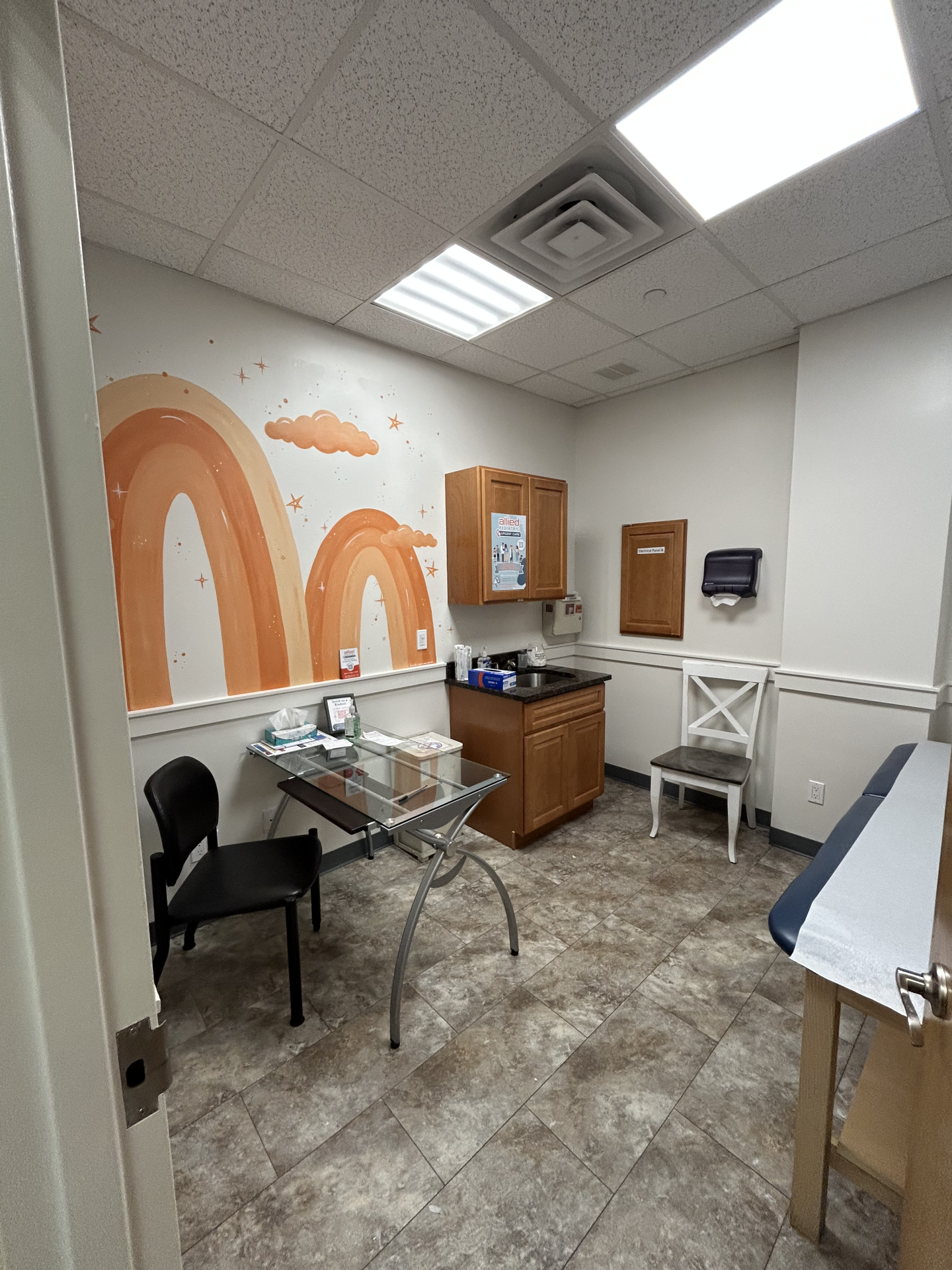 A doctor’s examination room with a mural of orange rainbows and stars on the wall, a glass desk with a black chair, and an examination table.