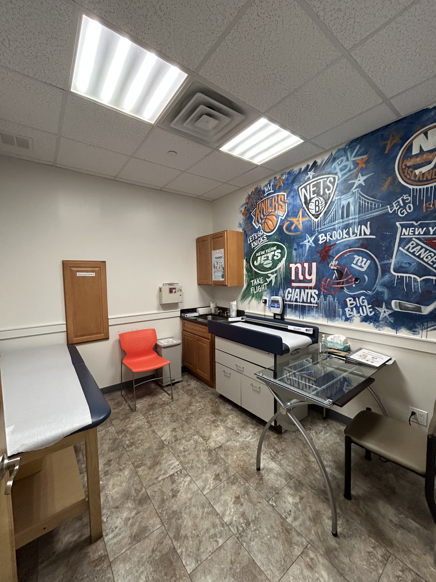 Examination room with a wall mural featuring New York sports team logos, a medical exam table, an orange plastic chair, and a desk with a computer.