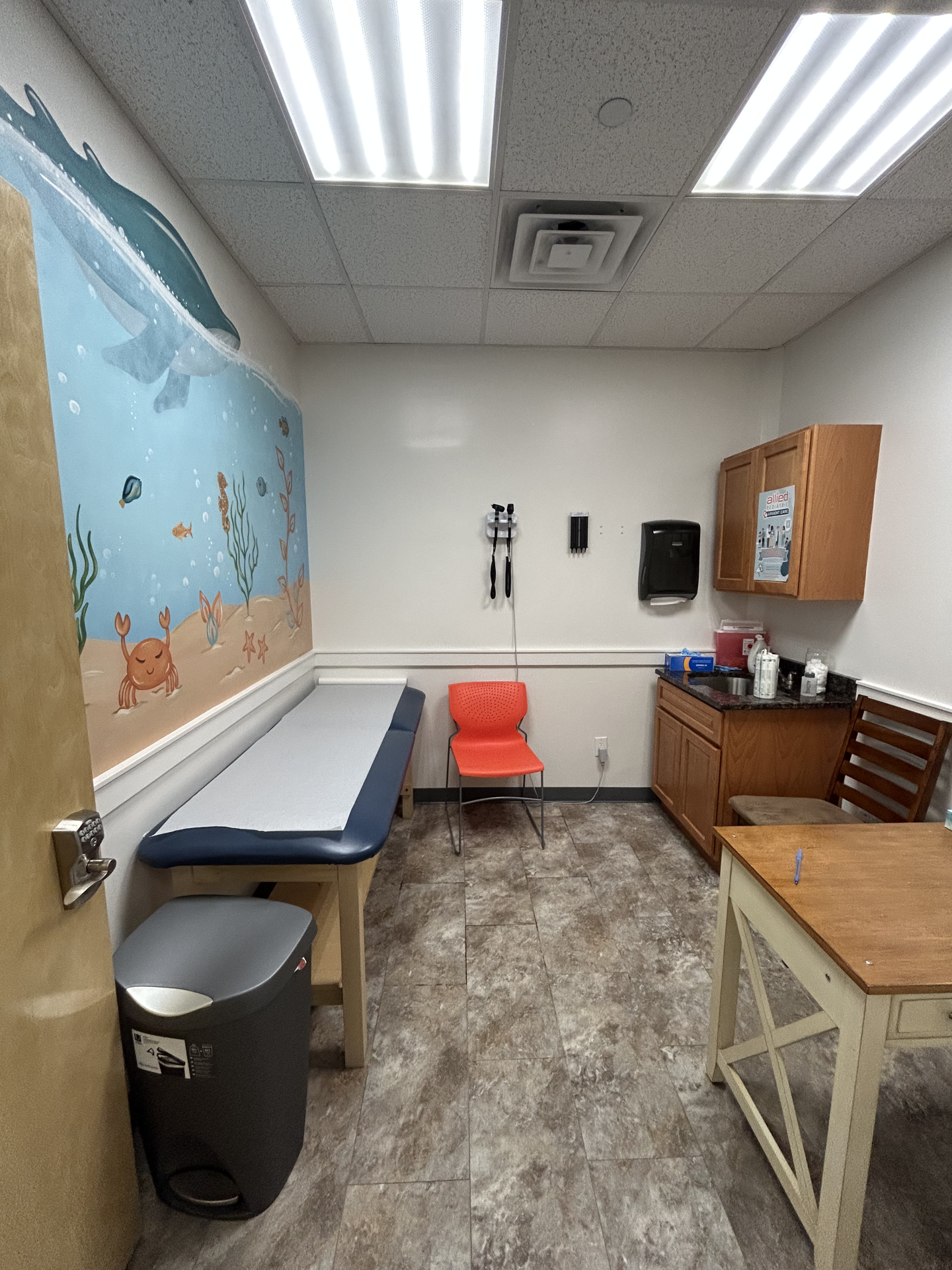 A pediatric examination room featuring an exam table and an orange chair, with an underwater-themed mural on the wall depicting a whale, crabs, and fish.