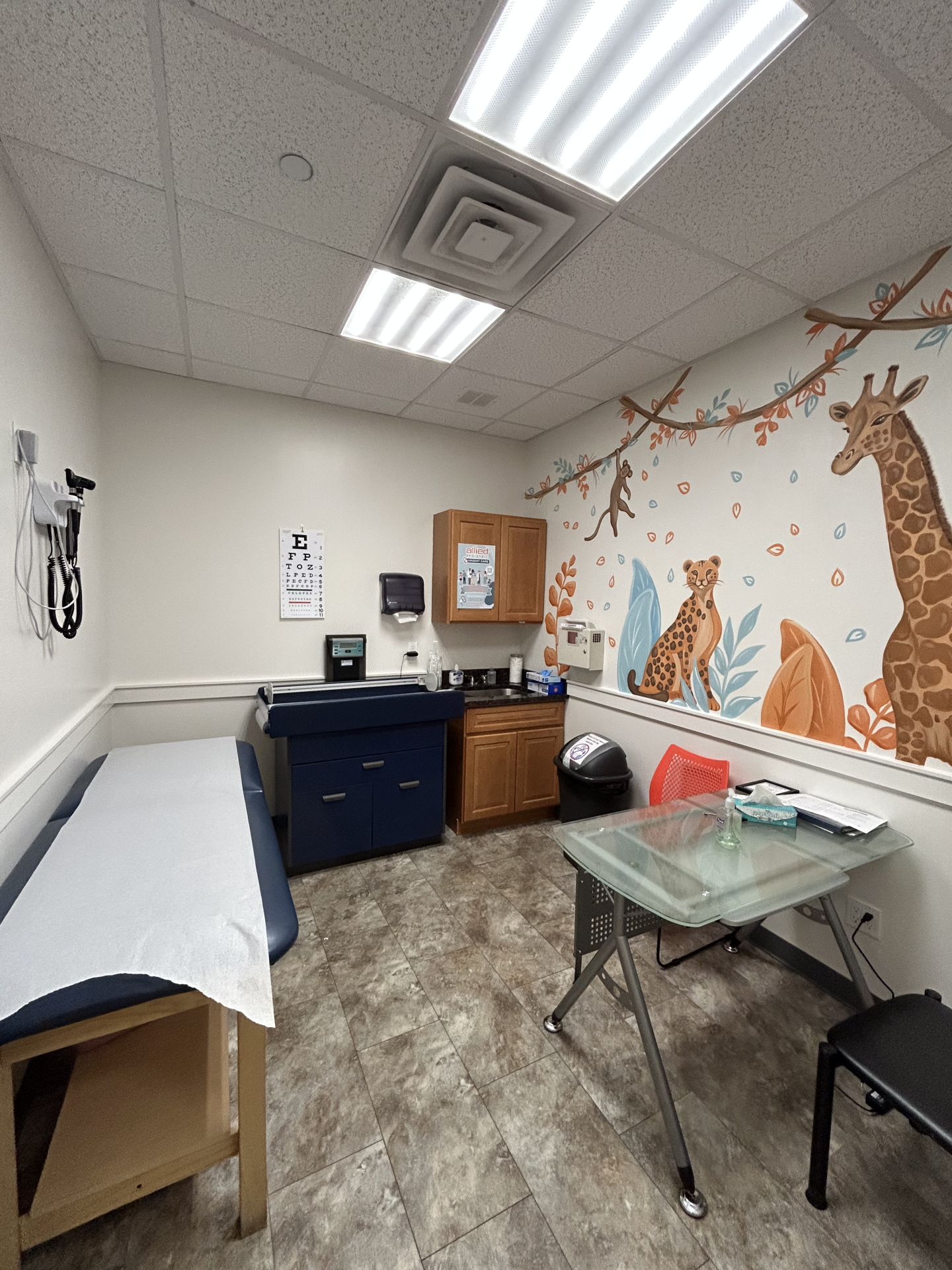 A pediatric examination room featuring a mural of a giraffe and leopard on the ceiling, a blue medical exam table, and an eye chart on the wall.