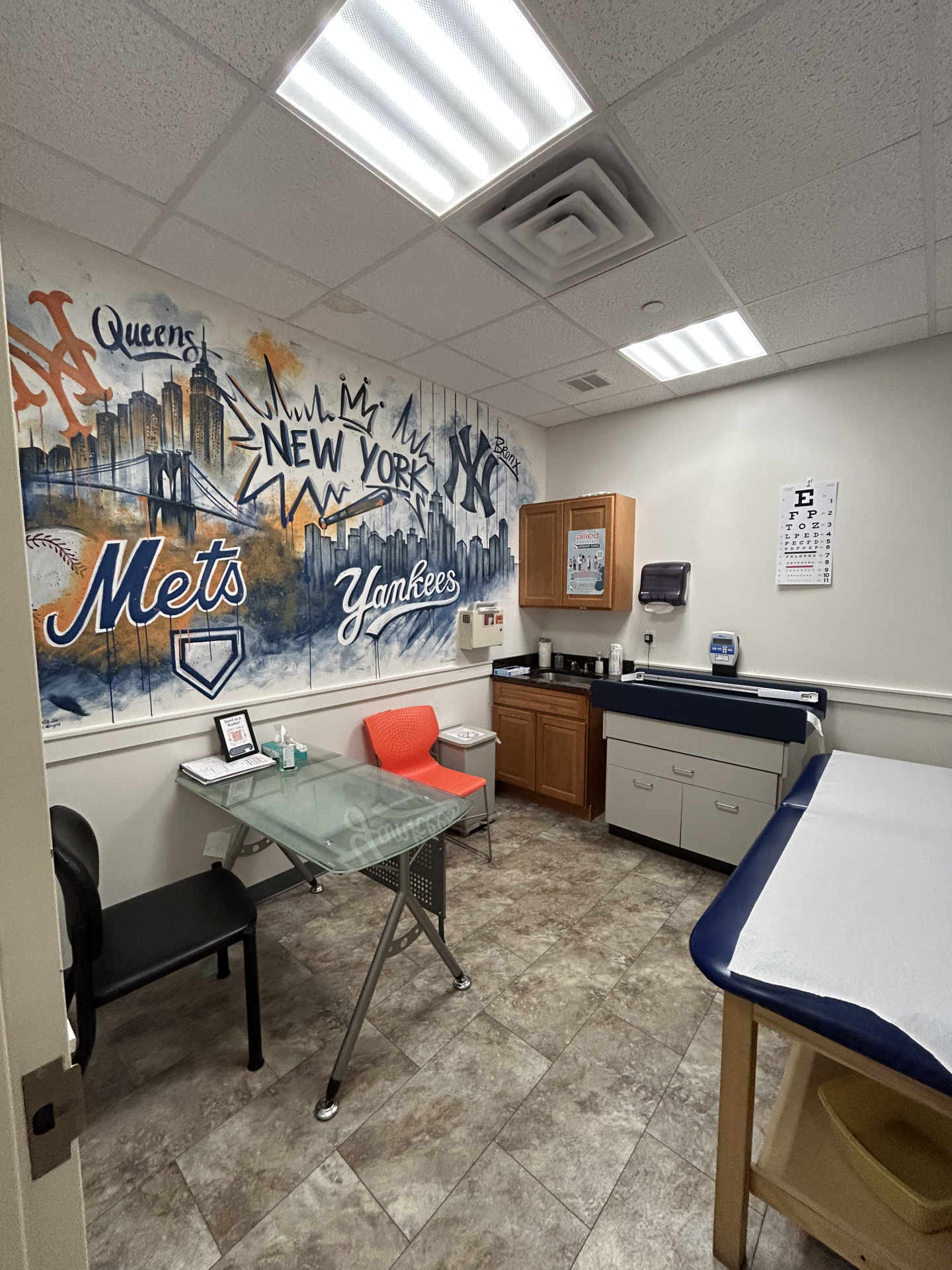 A medical examination room featuring a large wall mural of the New York Yankees and New York Mets logos, an exam table, a desk with two chairs, and a vision chart on the wall.