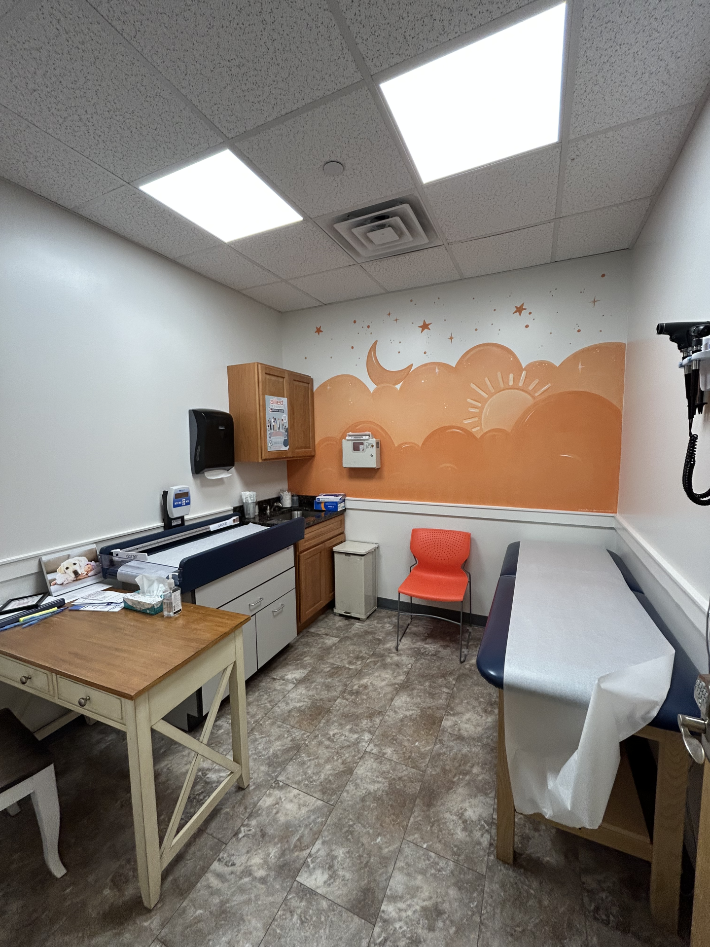 A medical examination room with an orange mural of clouds and stars, featuring an exam table with paper covering, a desk, and a wall-mounted supply dispenser.