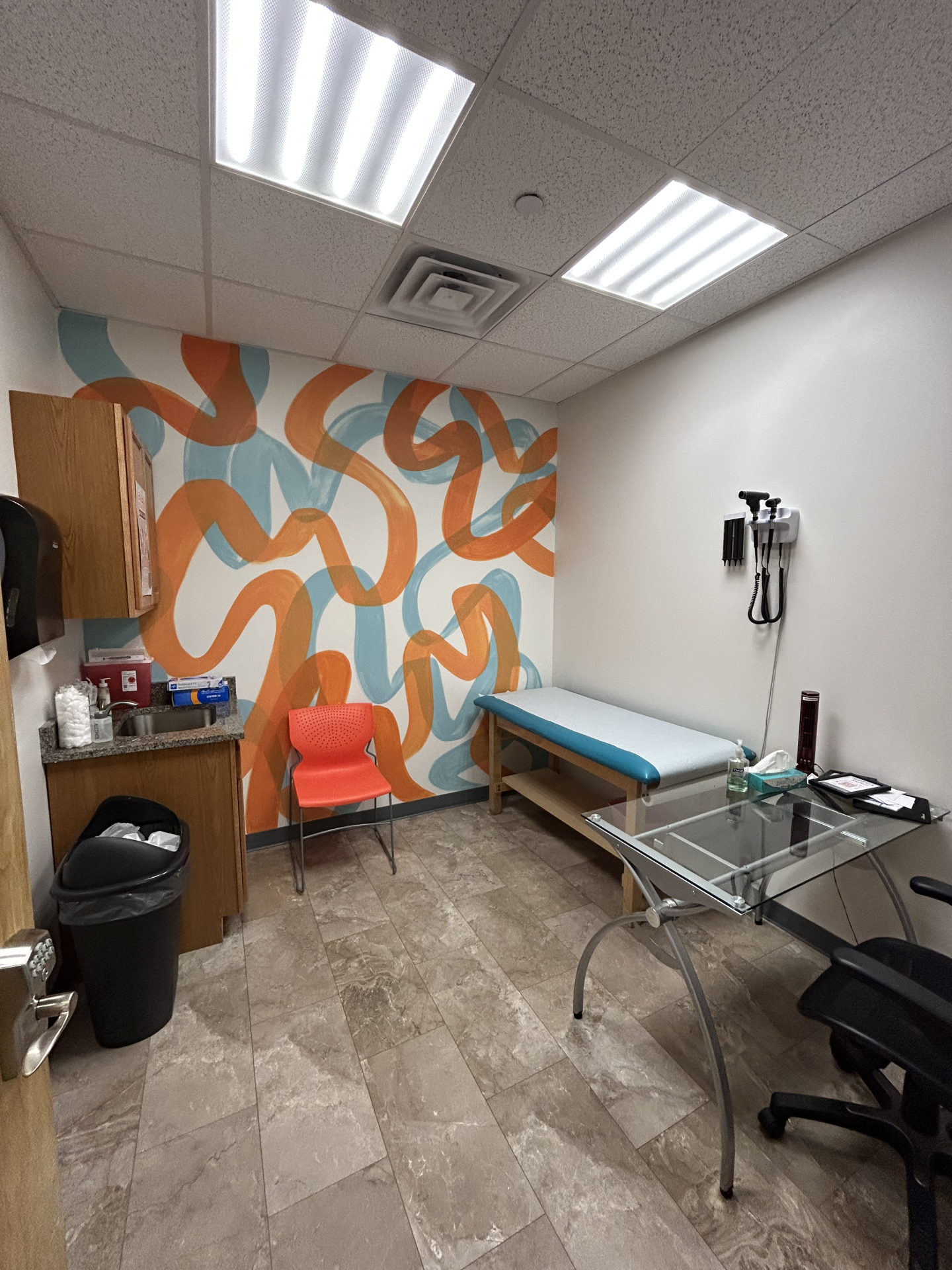 A medical exam room featuring a blue exam table, an orange chair, and an abstract orange and blue wall mural.