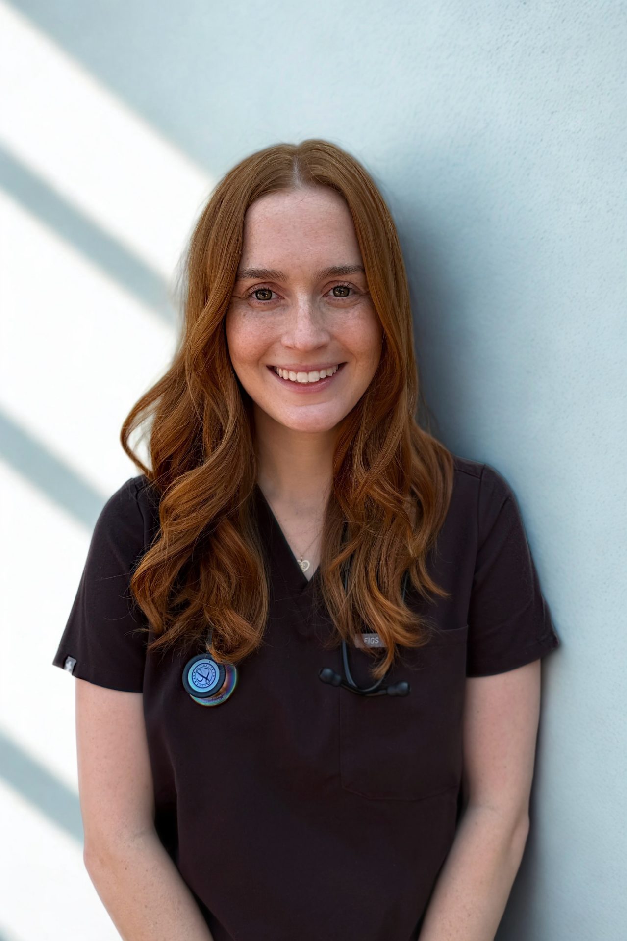 A smiling woman with long red hair, dressed in black medical scrubs and a stethoscope, stands in front of a light-colored wall with diagonal shadows.
