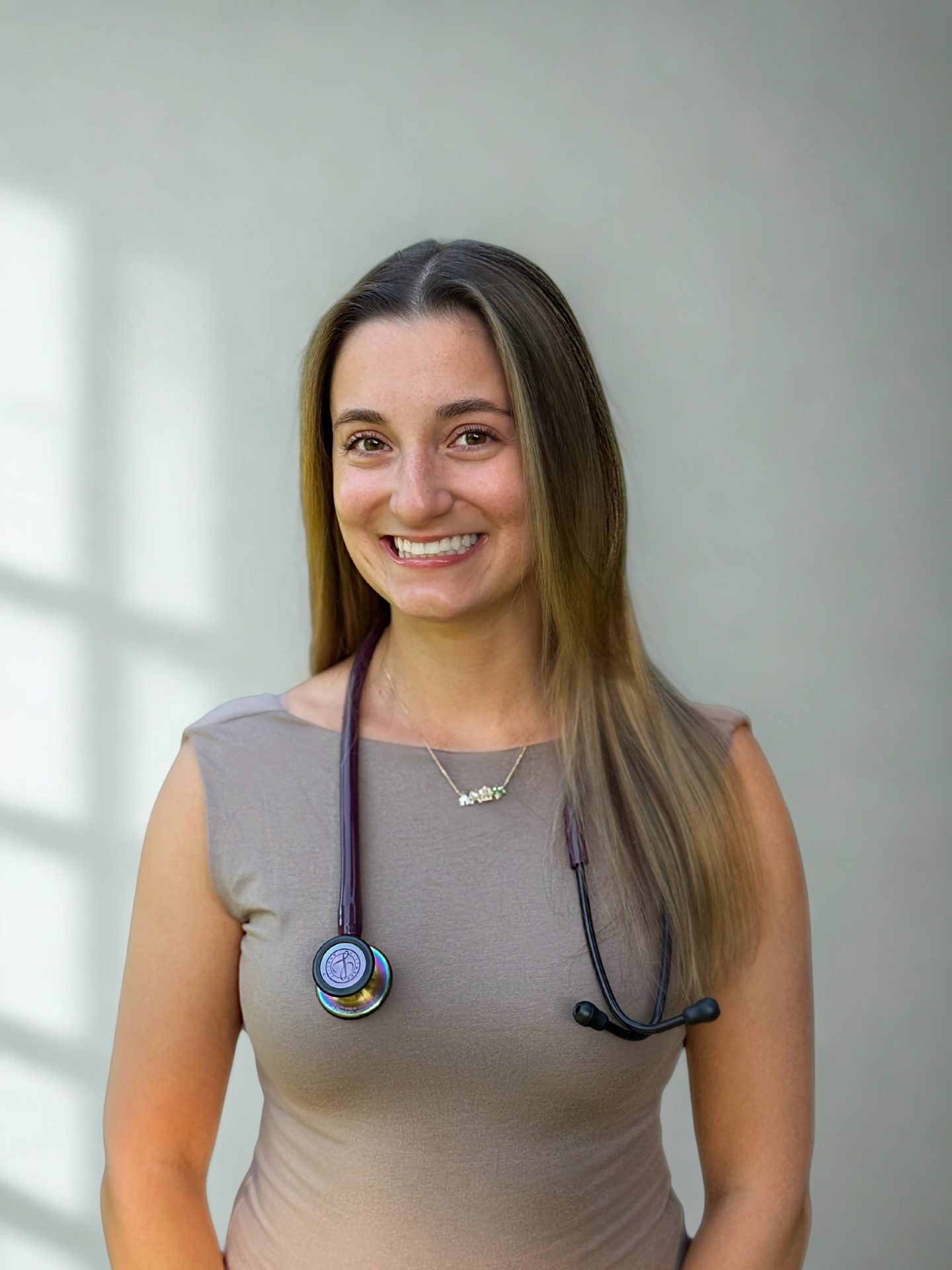 A woman with long brown hair, dressed in a sleeveless beige top and a stethoscope around her neck, smiles at the camera against a plain, softly lit background.