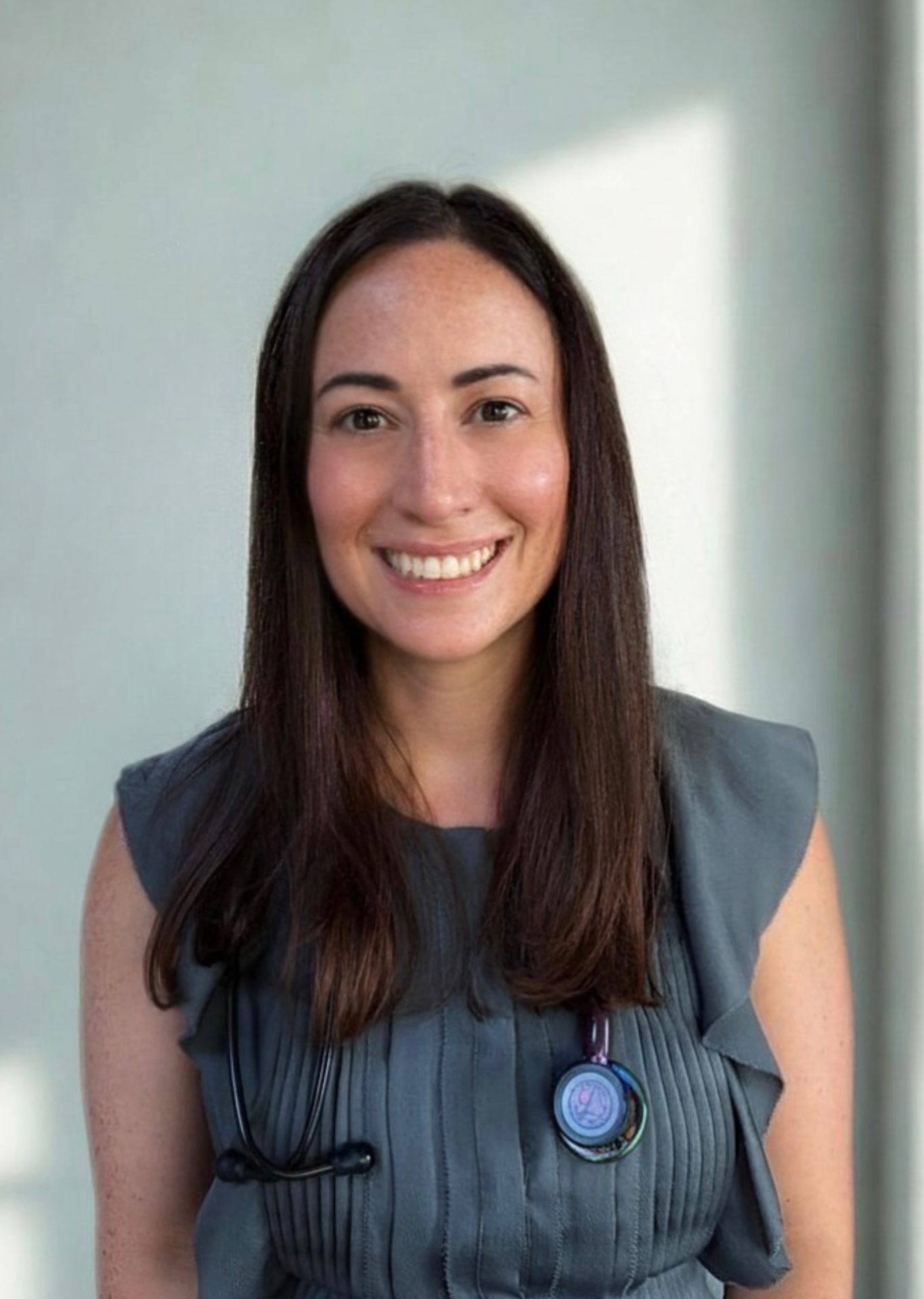 A woman with long brown hair, dressed in a gray top and a stethoscope around her neck, smiles at the camera in front of a light background.