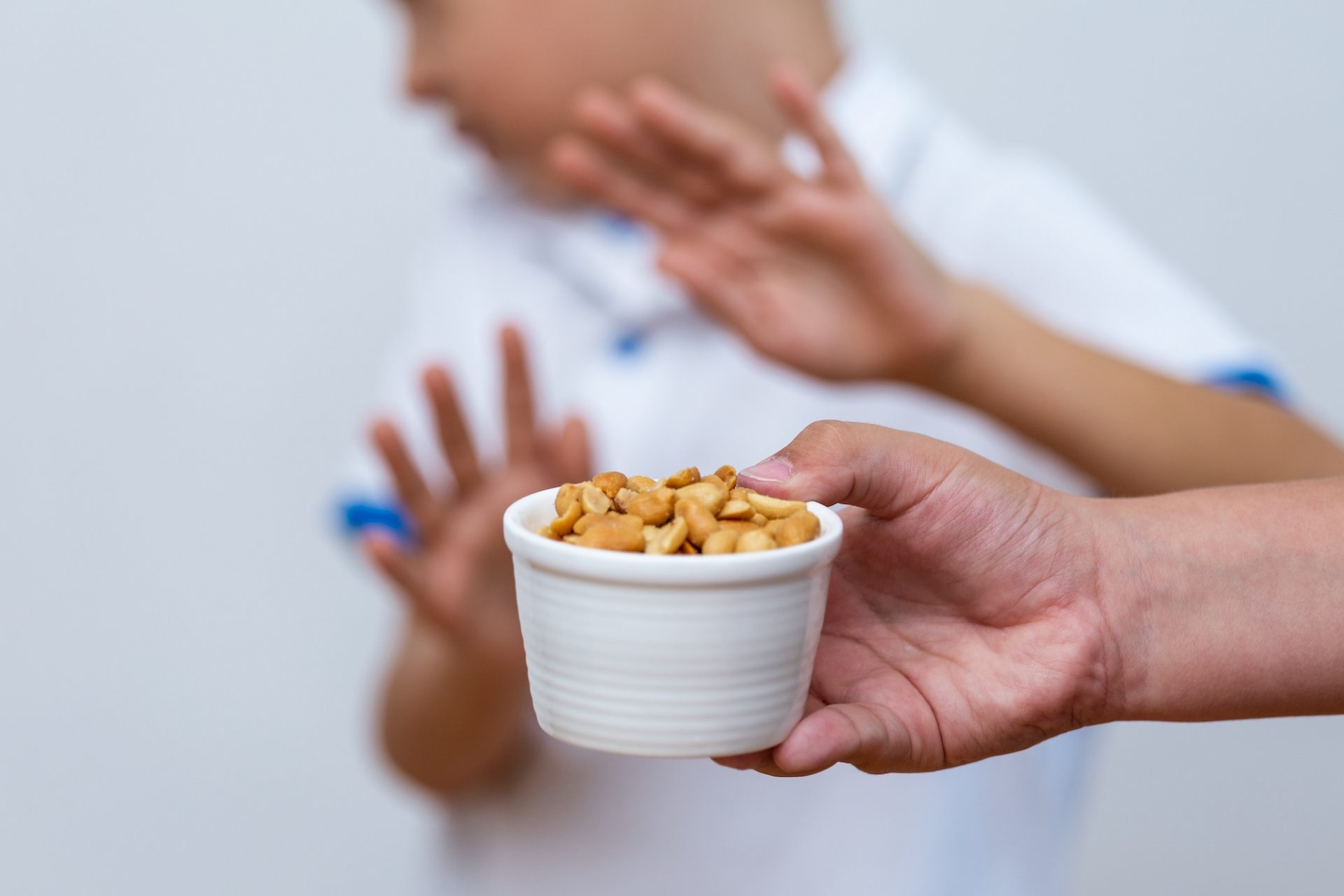 A person holds a bowl of peanuts, and in the background, a child turns away with raised hands, seemingly refusing or avoiding the food.