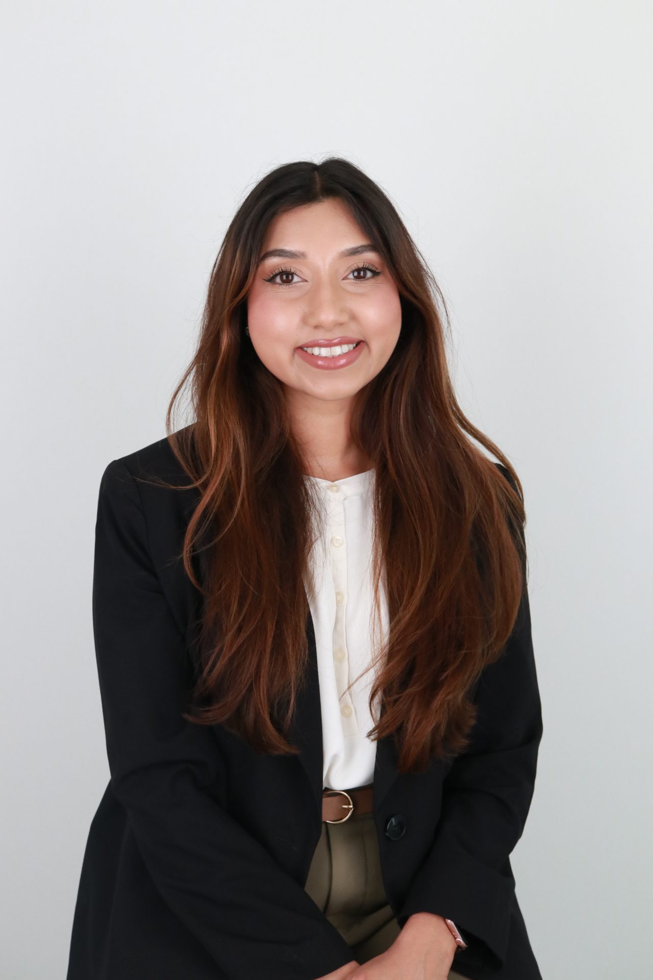 A woman with long brown hair, dressed in a black blazer over a white blouse, smiles while sitting in front of a simple light background.