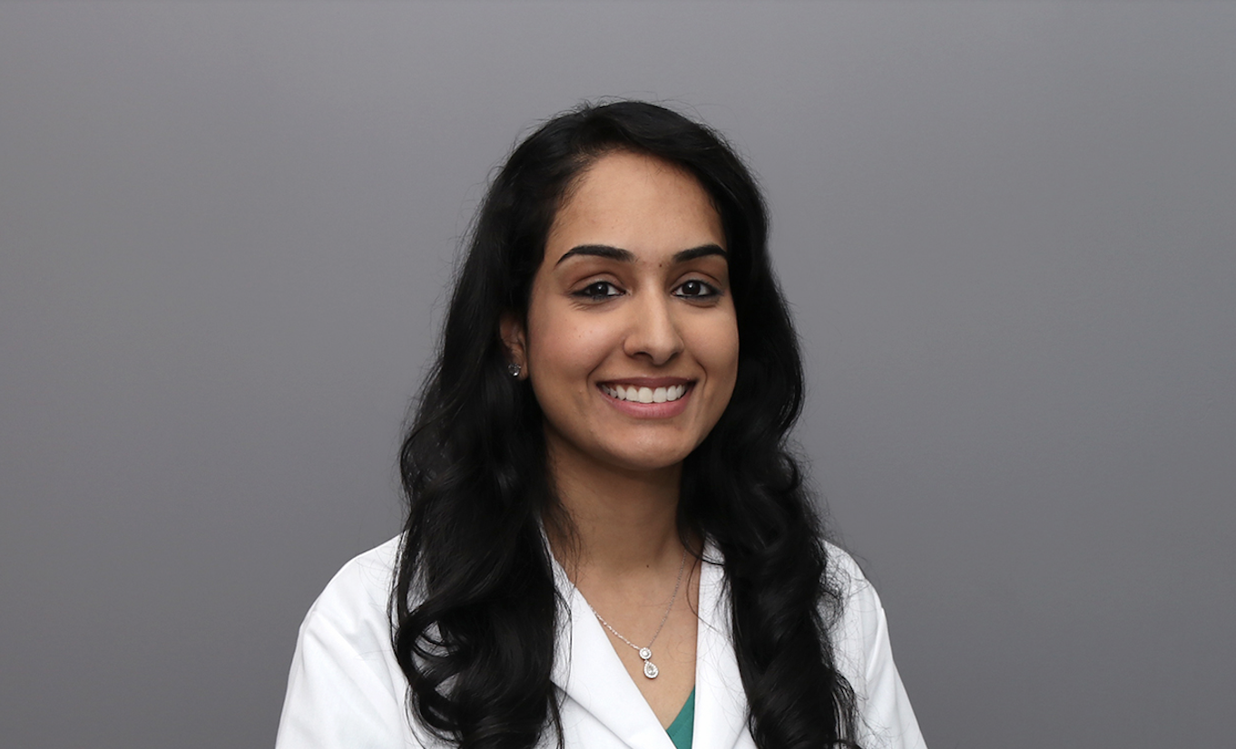 A woman with long dark hair, dressed in a white lab coat and a necklace, smiles as she stands before a plain gray background.