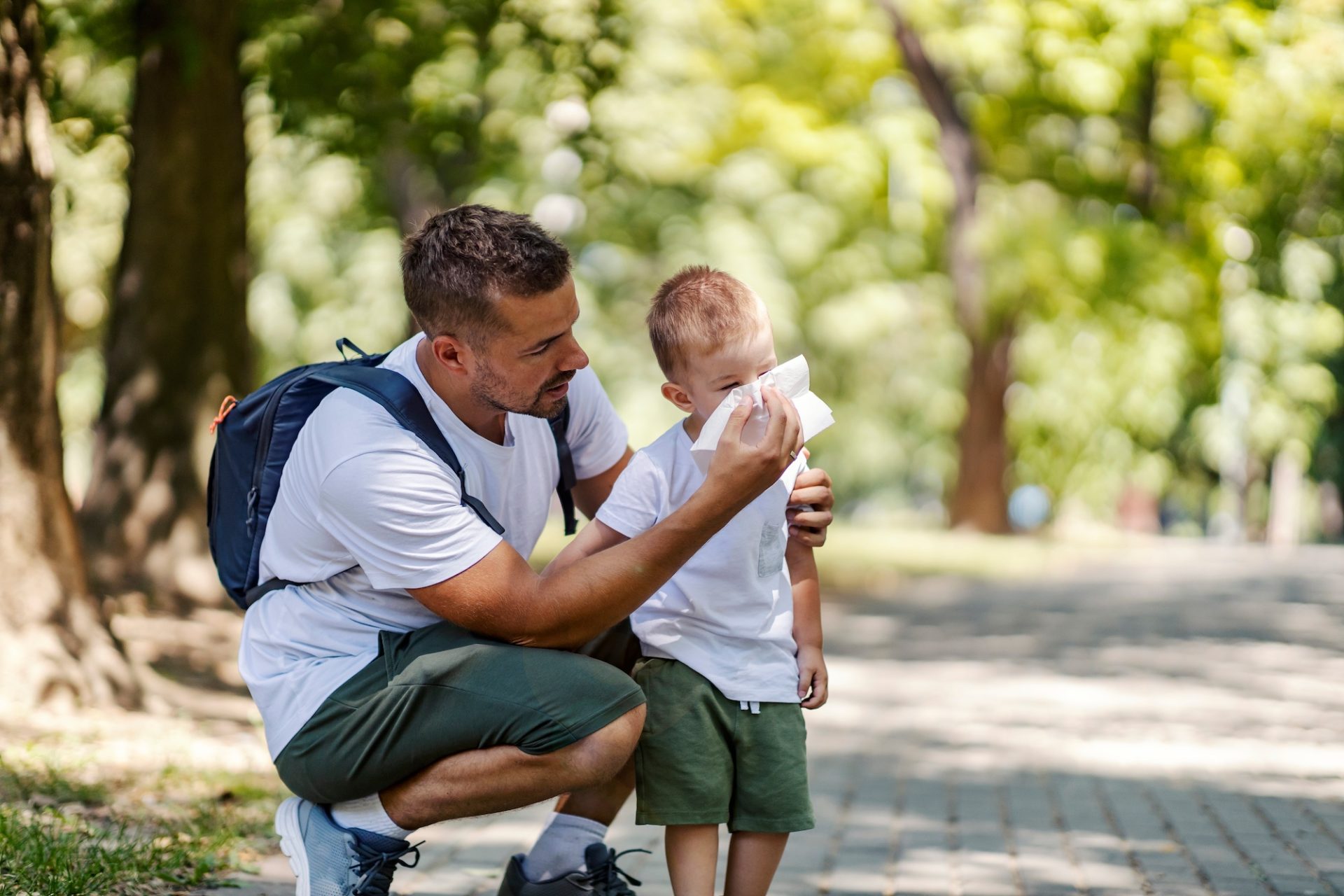 A man kneels in a park, gently wiping a young boy’s face with a tissue. Both wear white shirts and green shorts. Trees and sunlight fill the background.