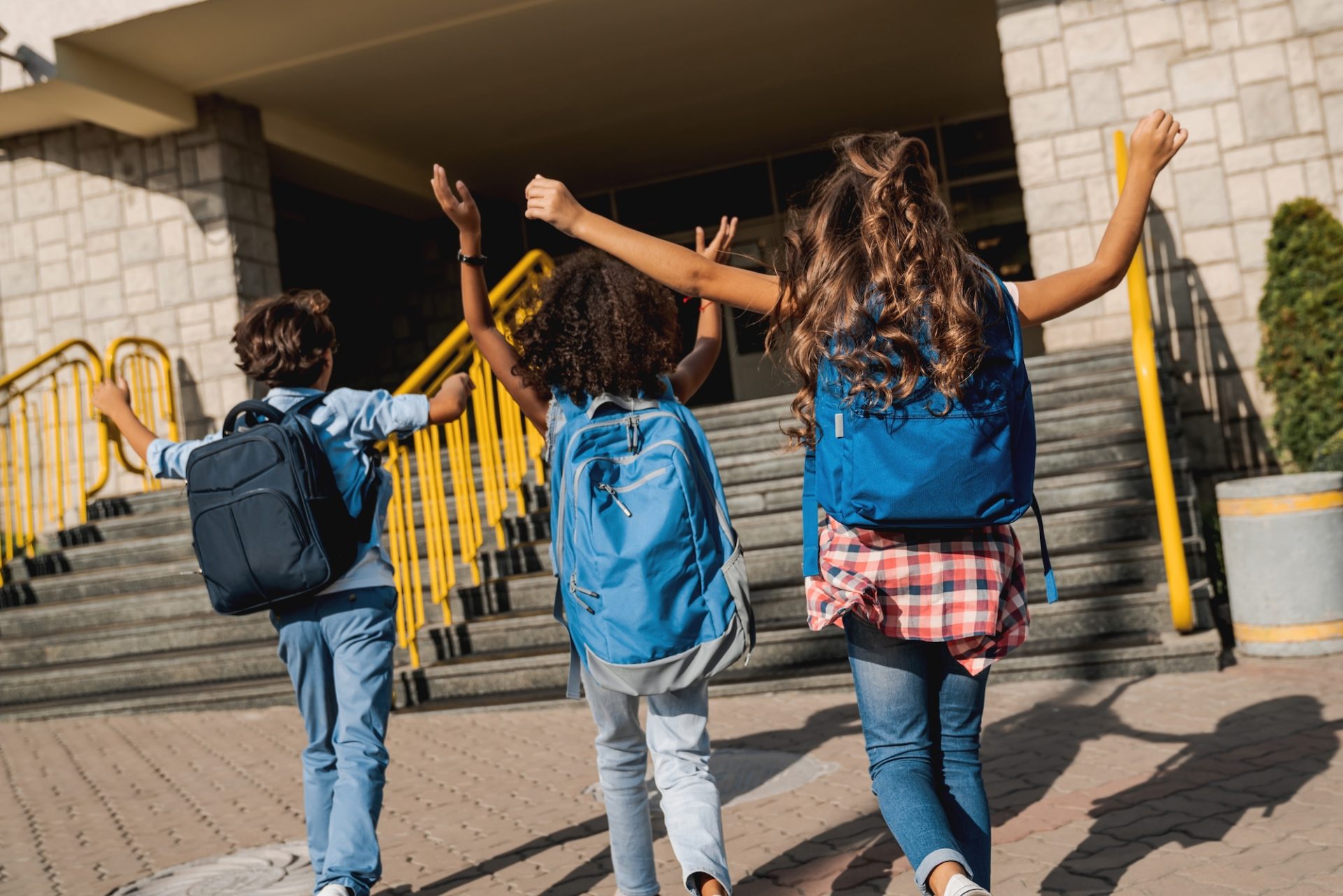 Three children with backpacks, seen from behind, walk joyfully toward a building with stairs and yellow railings, raising their arms in excitement, possibly arriving at or leaving school on a sunny day.