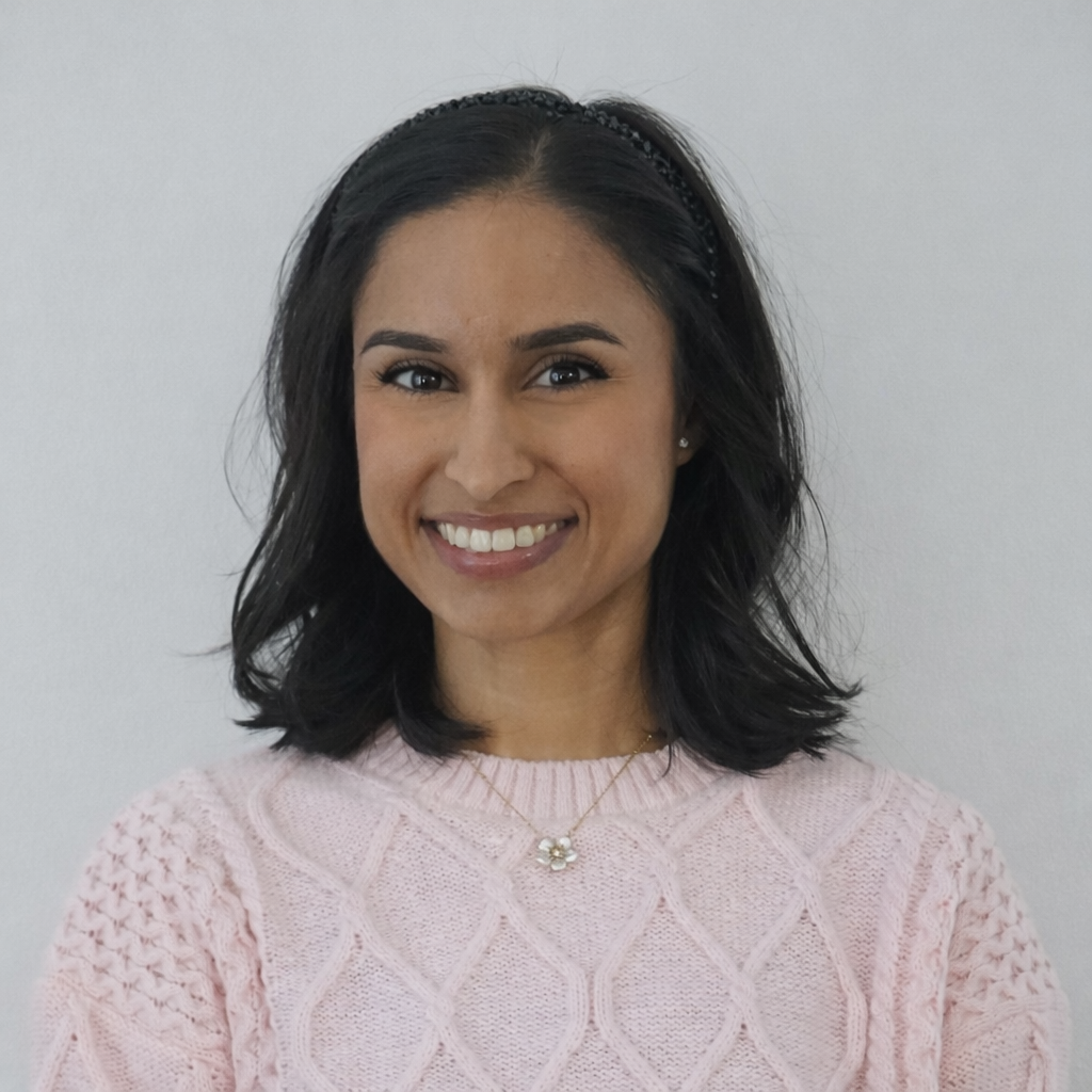 A smiling woman with dark hair wearing a light pink cable-knit sweater and a flower pendant necklace.